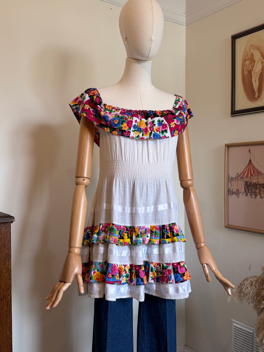 A white smocked dress displayed on a wooden mannequin, featuring colorful floral ruffle trim at the off-the-shoulder neckline and the hem. The dress is styled with blue jeans on a mannequin in an indoor room setting.