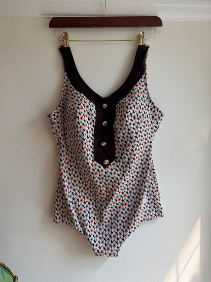 A sleeveless water swimsuit with a dense small-scale multicolor geometric pattern on a white base, featuring a solid black V-neckline panel with three silver snap buttons, hanging on a wooden hanger against a plain white wall.