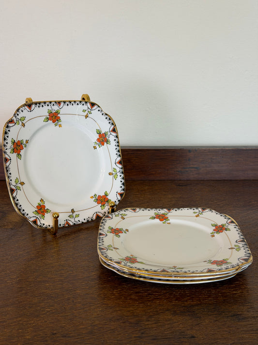 A set of vintage square ceramic plates with a white base, decorated with a floral border pattern in orange, green, and gold, displayed on a dark wooden table against a plain wall. Set of four vintage Roslyn China R & Co England square plates with orange floral sprays, green leaves, geometric black and burnt-orange borders and gold detailing.