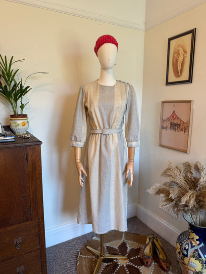
Vintage neutral beige midi dress with a belted waist, soft long sleeves and minimalist panelled design displayed on a mannequin with a red headscarf.