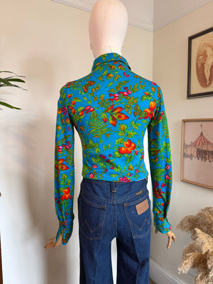 A mannequin wearing a bright turquoise vintage 1970s long-sleeve button-up shirt with a dense multicolor floral and butterfly print, paired with dark blue jeans, shown indoors against a cream wall.