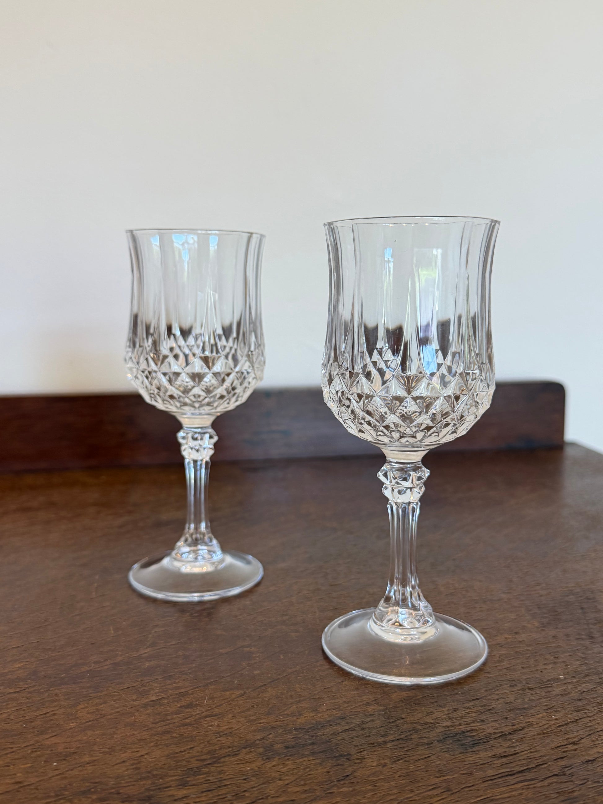 Two clear cut-crystal wine glasses with faceted stems and round bases, displayed on a dark wooden table against a plain off-white wall.
