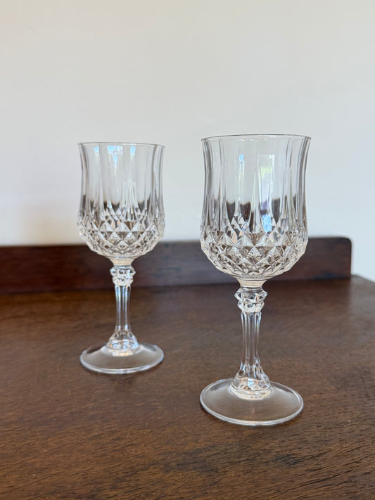 Two clear cut-crystal wine glasses with faceted stems and round bases, displayed on a dark wooden table against a plain off-white wall.