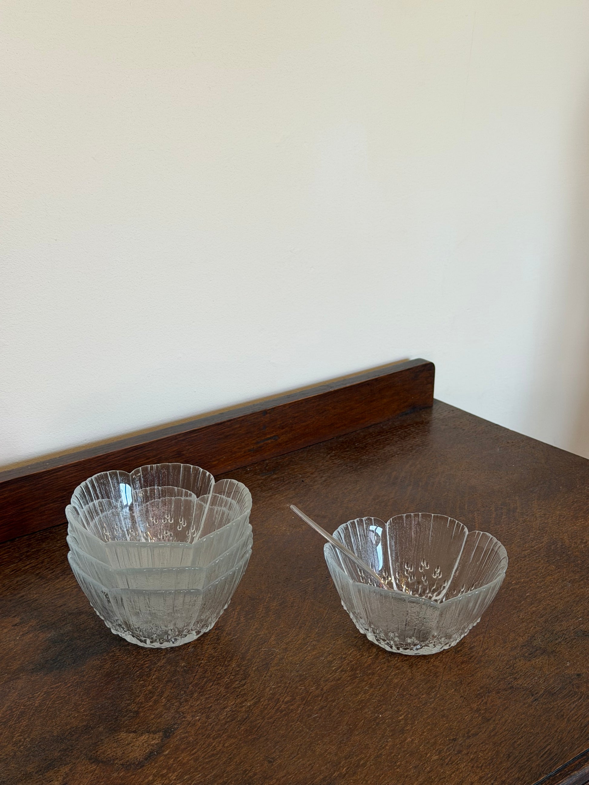Set of four vintage frosted glass dessert bowls with flower-shaped scalloped edges and textured pressed glass detail, photographed on a dark wooden surface against an off-white wall.