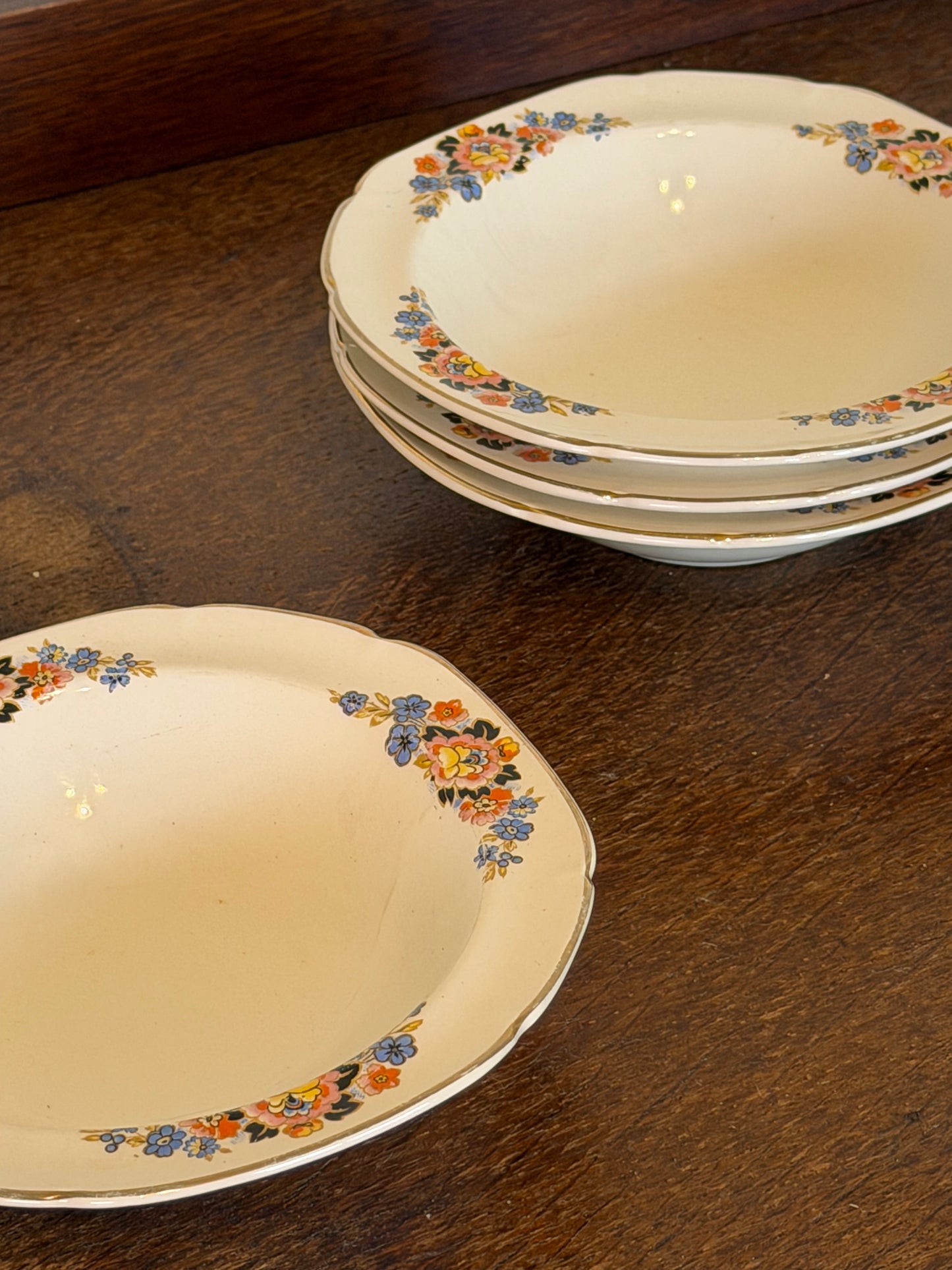 Set of four vintage John Maddock & Sons Ivory Ware shallow soup bowls dating from the 1940s–1950s, featuring a cream ground, colourful floral border in blue, pink and orange, and fine gold edging, arranged on a dark wooden surface.
