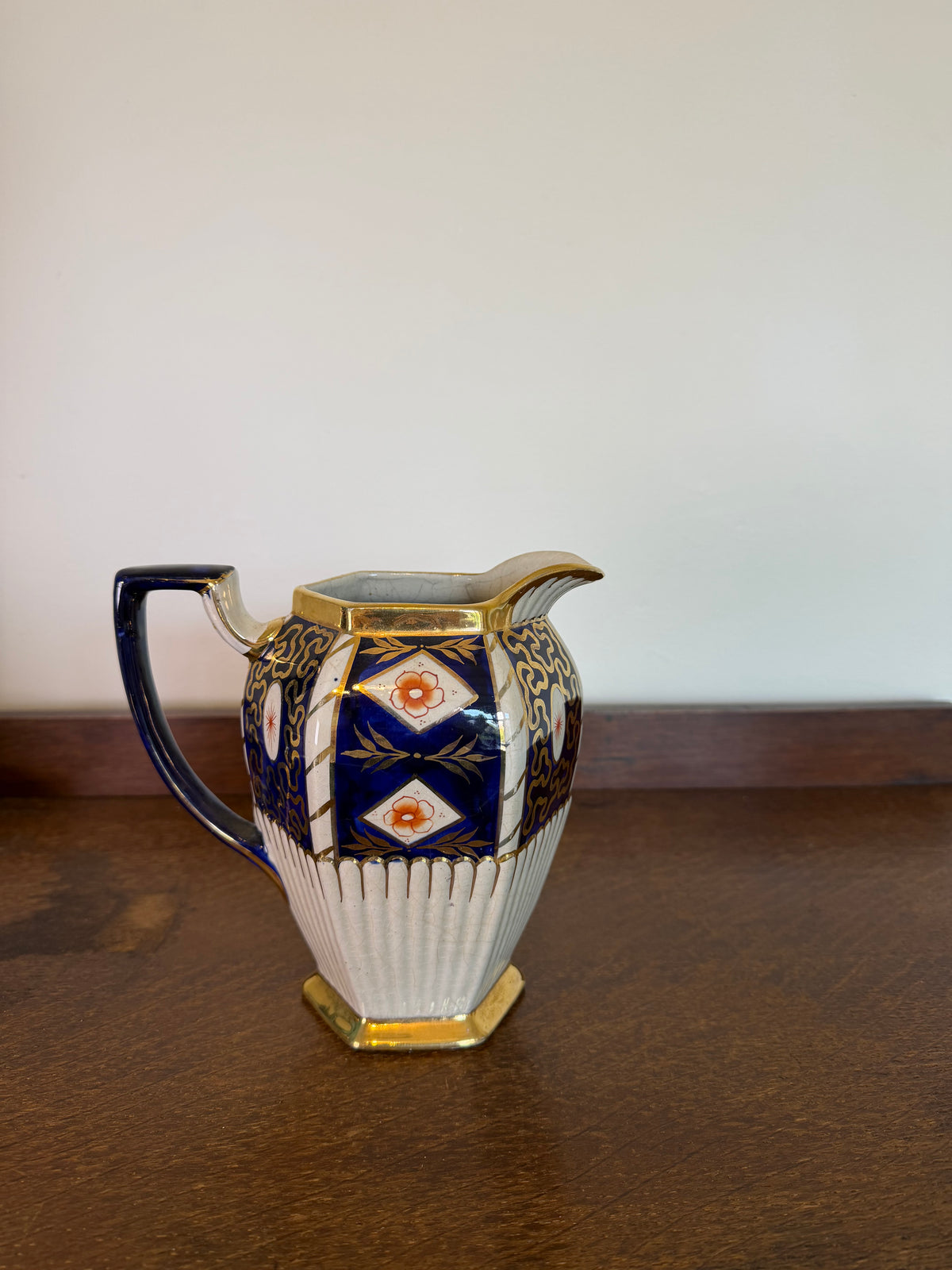 A vintage octagonal ceramic creamer pitcher with a blue and gold geometric pattern and a ribbed white lower body, sitting on a dark wooden table against a plain off-white wall.