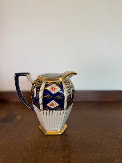 A vintage octagonal ceramic creamer pitcher with a blue and gold geometric pattern and a ribbed white lower body, sitting on a dark wooden table against a plain off-white wall.