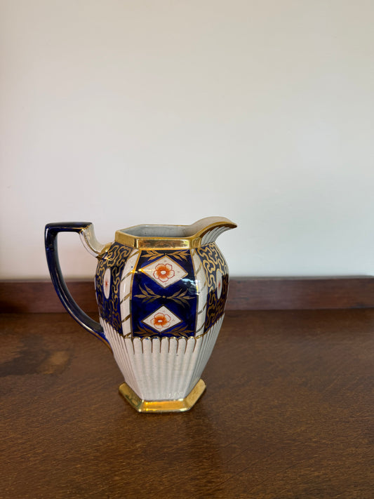 A vintage octagonal ceramic creamer pitcher with a blue and gold geometric pattern and a ribbed white lower body, sitting on a dark wooden table against a plain off-white wall.