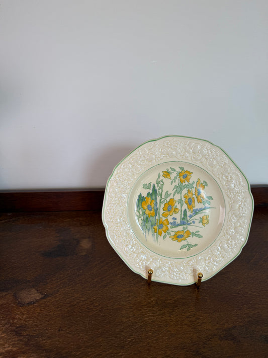 A vintage ceramic wall plate with a yellow and green floral design, mounted on a dark wooden wall next to a white wall.
Vintage Crown Ducal Florentine floral decorative plate with embossed border and yellow flower illustration.