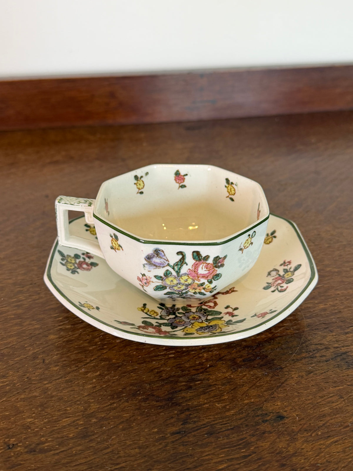 An octagonal ceramic teacup and saucer set with a delicate multicolor floral pattern, resting on a dark wooden table.