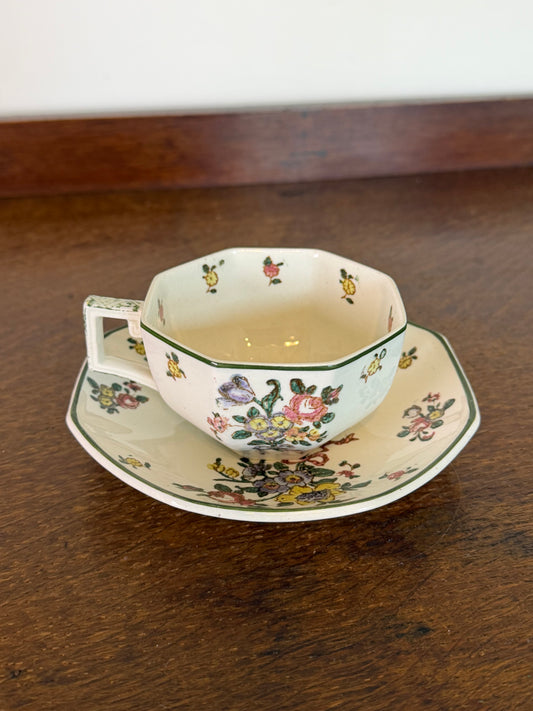 An octagonal ceramic teacup and saucer set with a delicate multicolor floral pattern, resting on a dark wooden table.