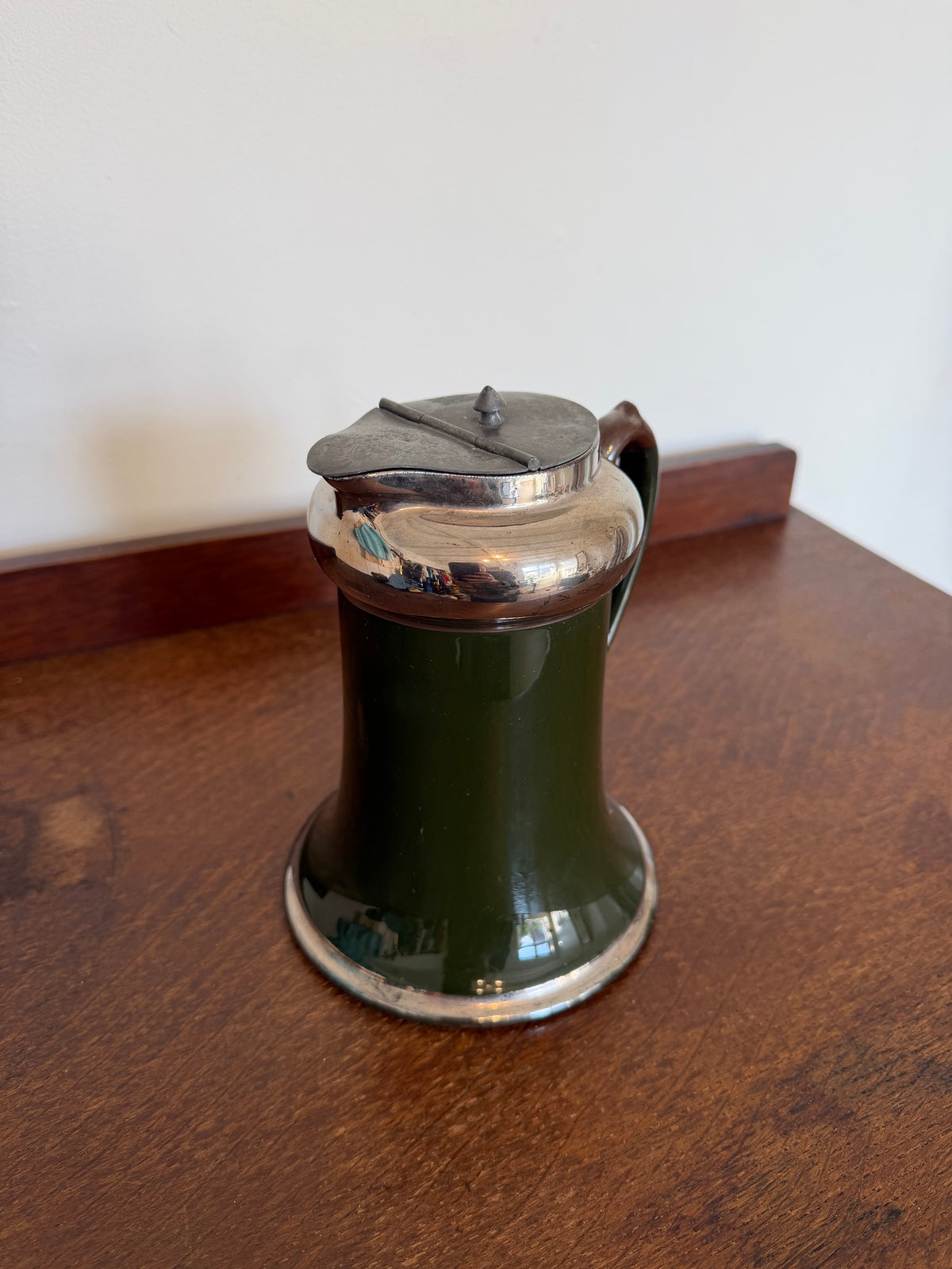 A vintage dark green ceramic pitcher with a polished silver-tone metal lid and handle, sitting on a wooden table against a plain wall.