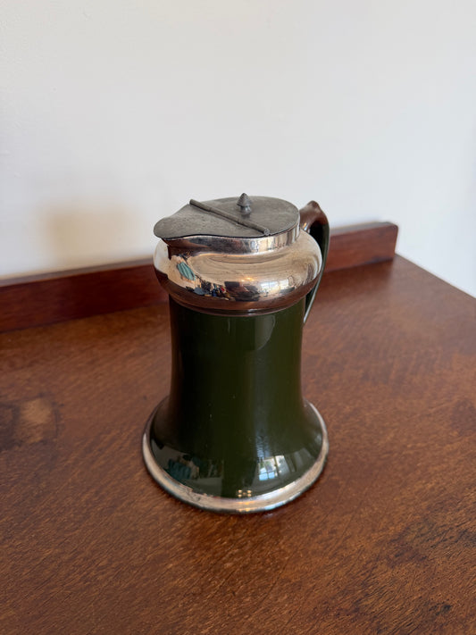 A vintage dark green ceramic pitcher with a polished silver-tone metal lid and handle, sitting on a wooden table against a plain wall.
