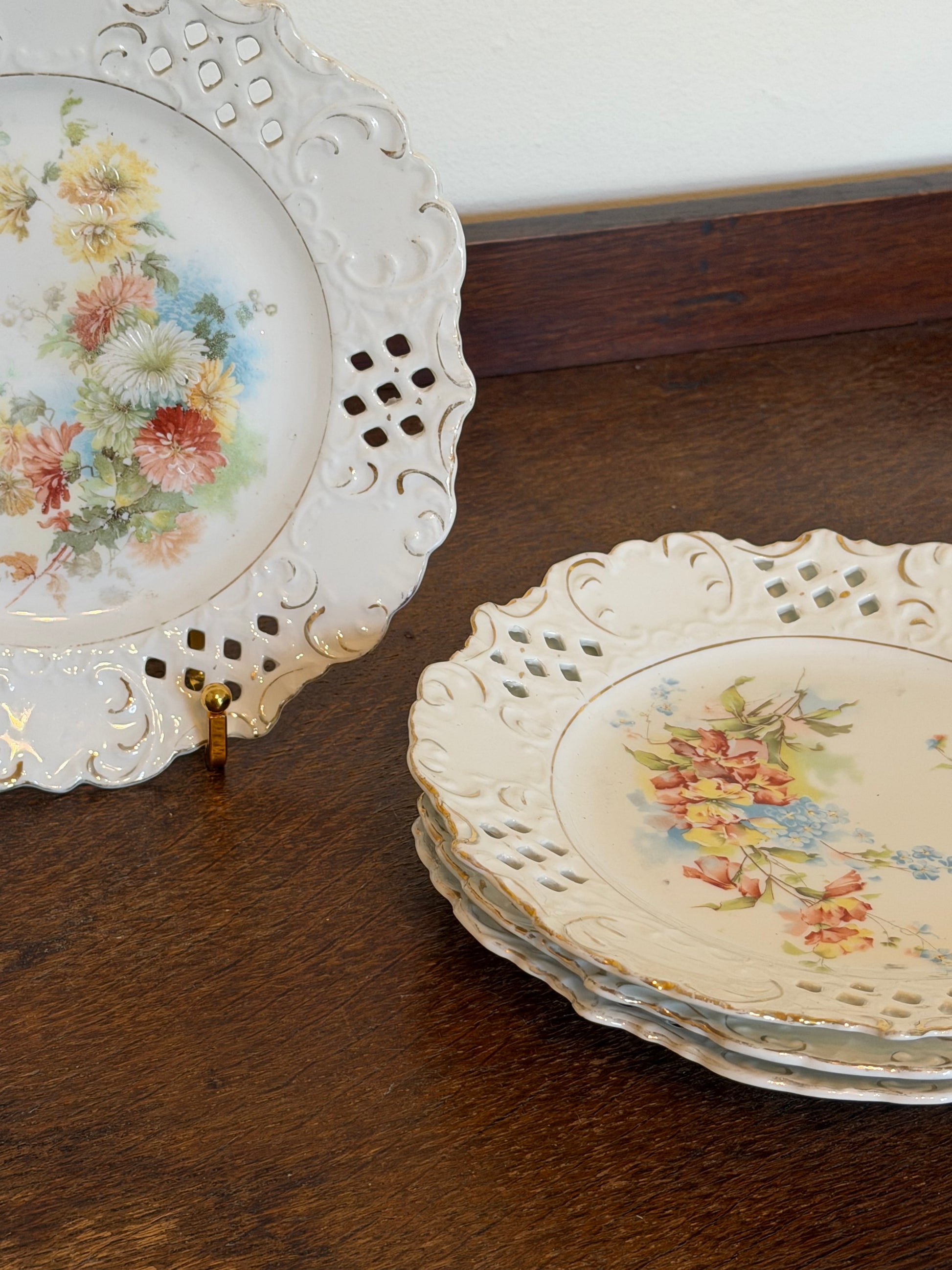 Set of four vintage porcelain dessert plates dating from the 1930s–1950s, featuring pastel floral centres, ornate pierced borders with raised scrollwork and gold detailing, arranged on a dark wooden surface.