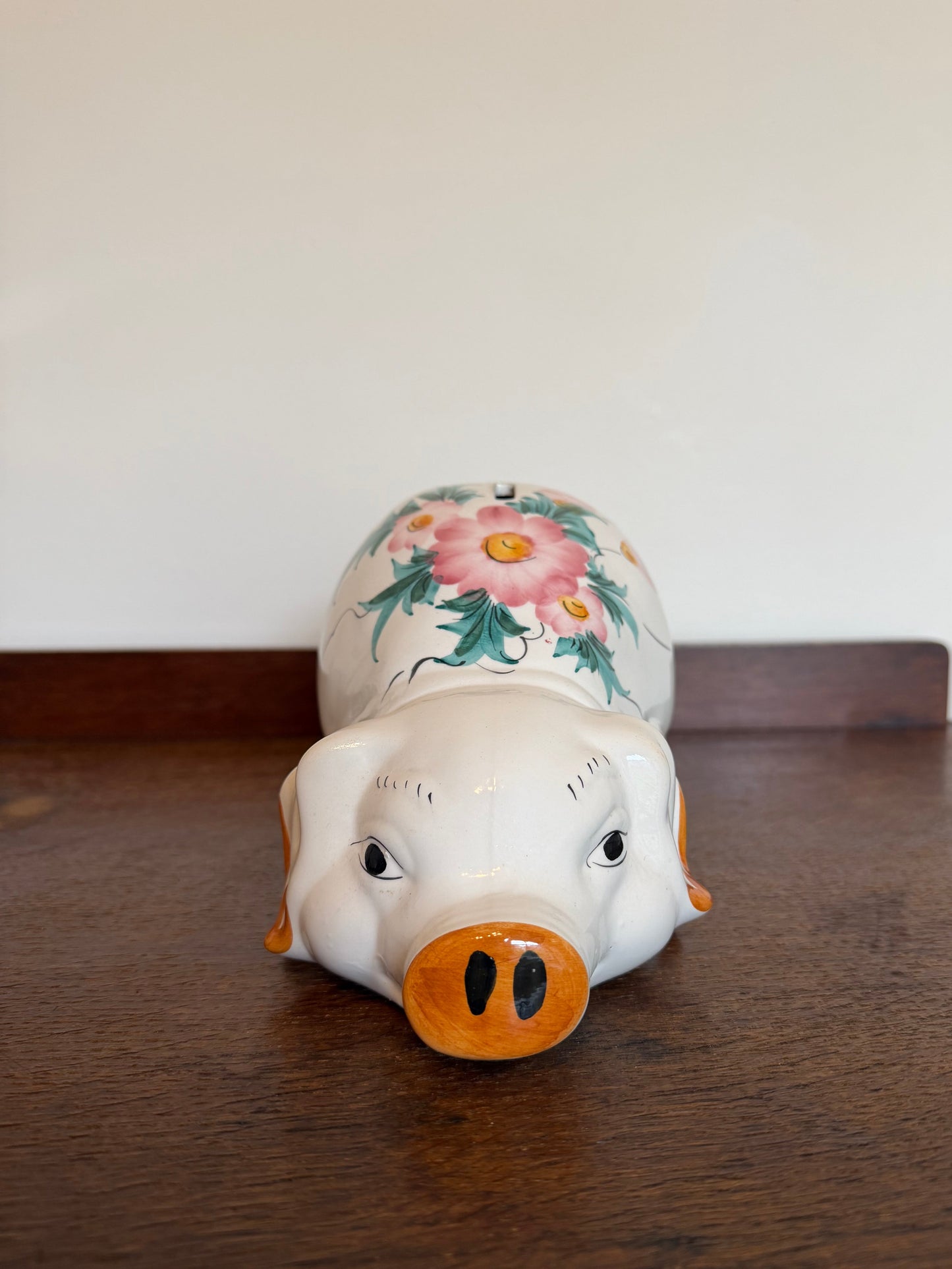 A ceramic pig-shaped pig bank or figurine lying on its side on a dark wooden table against a plain off-white wall. The pig has a white body, a bright orange snout and ears, and a floral design painted on its back with pink flowers and green leaves.