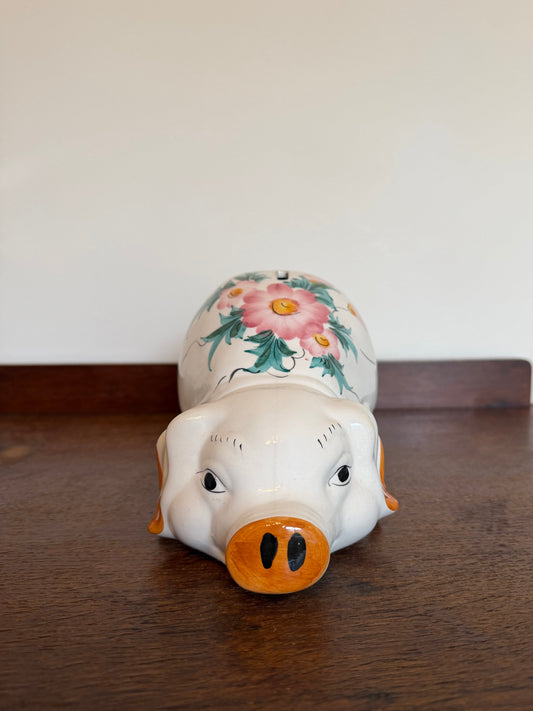 A ceramic pig-shaped pig bank or figurine lying on its side on a dark wooden table against a plain off-white wall. The pig has a white body, a bright orange snout and ears, and a floral design painted on its back with pink flowers and green leaves.