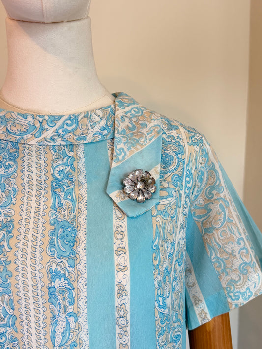 A close-up of a light blue and beige patterned blouse displayed on a mannequin. The blouse features vertical stripes with ornate paisley and chain-like patterns, a pointed collar, and a decorative silver-toned flower brooch pinned near the collar.