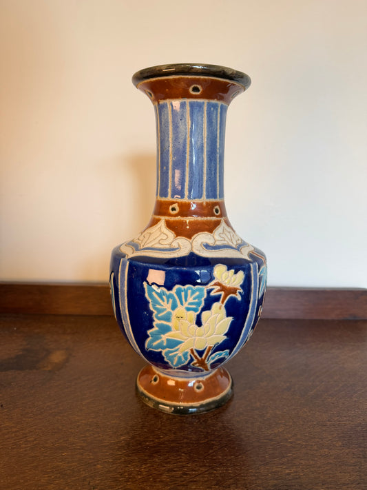 Vintage 1960s Japanese style ceramic vase in cobalt blue with hand painted floral and botanical decoration, brown glazed detailing and a tall flared neck.set on a dark wooden surface against a plain beige wall.
