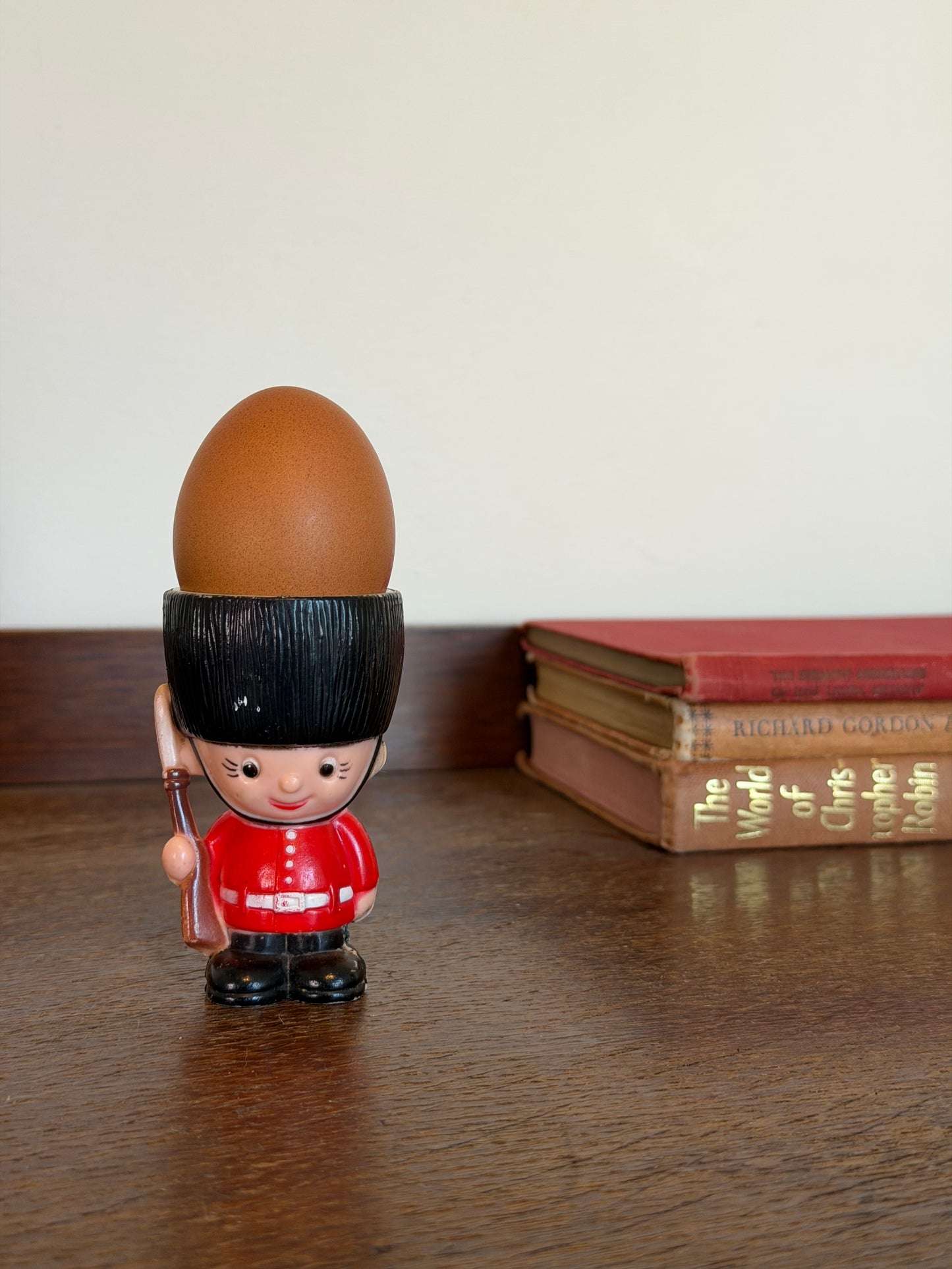 Vintage novelty egg cup shaped like a British Guardsman with a red uniform, black bearskin hat and small rifle, photographed on a dark wooden surface against a cream wall.