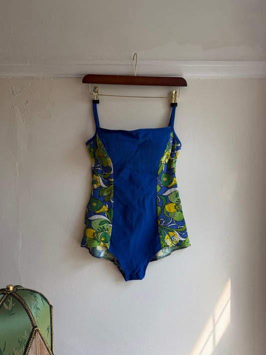 A blue one-piece swimsuit with green and yellow floral patterned trim, hanging from a wooden hanger against an off-white wall.