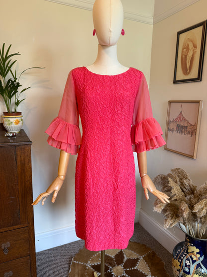 Vintage 1960s hot pink cocktail dress with a textured floral fabric, fitted wiggle silhouette and sheer sleeves finished with dramatic layered flounce cuffs, displayed on a mannequin.