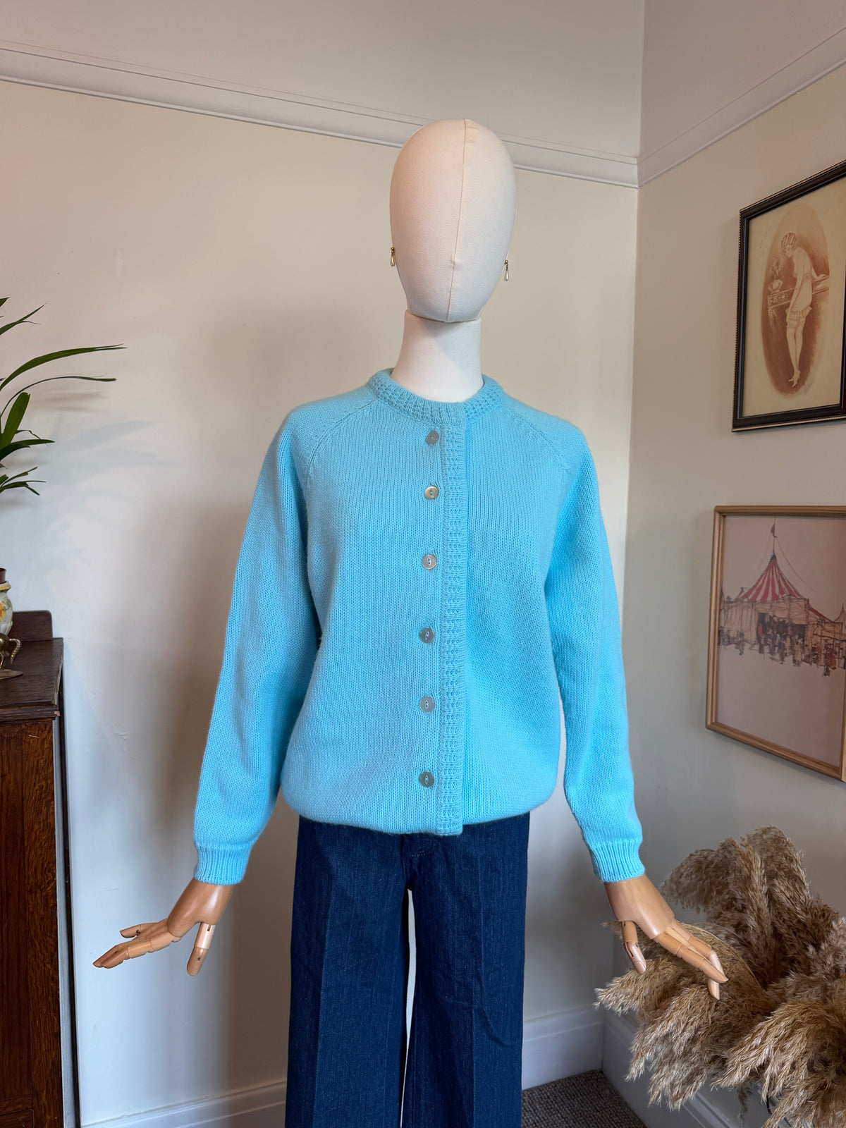A light blue knit cardigan sweater displayed on a mannequin, paired with dark blue wide-leg trousers, set against an indoor wall background with framed art and a plant.