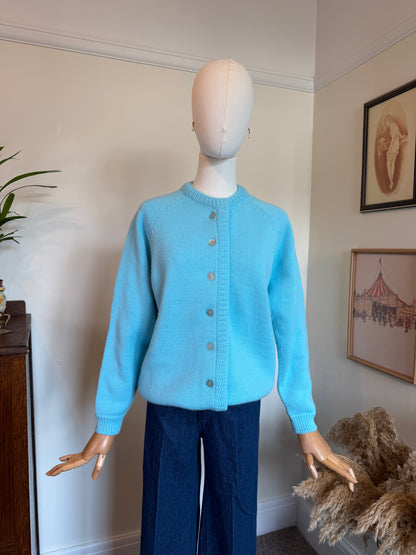A light blue knit cardigan sweater displayed on a mannequin, paired with dark blue wide-leg trousers, set against an indoor wall background with framed art and a plant.