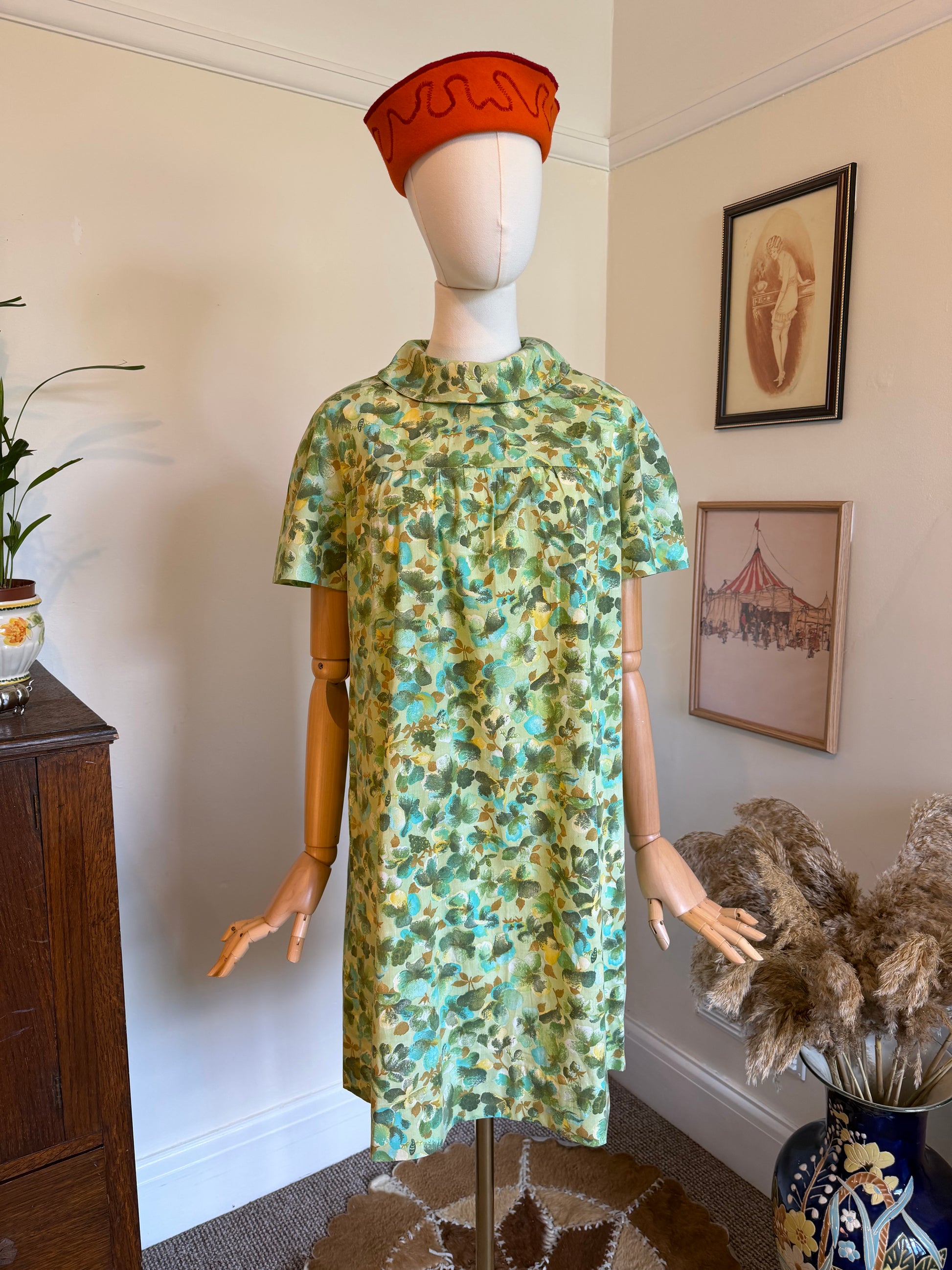 Vintage 1960s green floral shift dress with a painterly turquoise and olive botanical print, roll collar neckline and short sleeves, displayed on a mannequin.