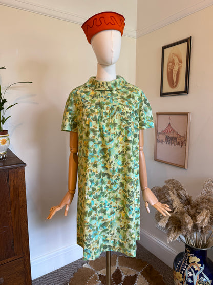 Vintage 1960s green floral shift dress with a painterly turquoise and olive botanical print, roll collar neckline and short sleeves, displayed on a mannequin.