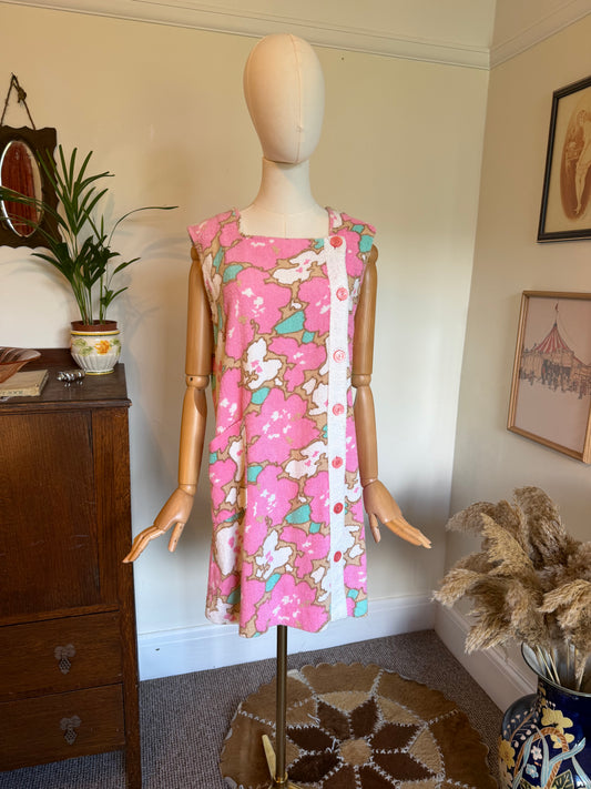 Vintage 1960s sleeveless mini dress in pink floral terrycloth fabric with an abstract flower print in pink, cream, tan and aqua, featuring a square neckline and decorative button-front panel, displayed on a mannequin.