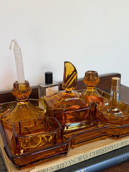 Vintage amber glass vanity set consisting of a large rectangular tray, lidded trinket box, two matching candlestick holders, a ring holder and small dish, styled with jewellery and perfume bottles on a dark wooden surface.
