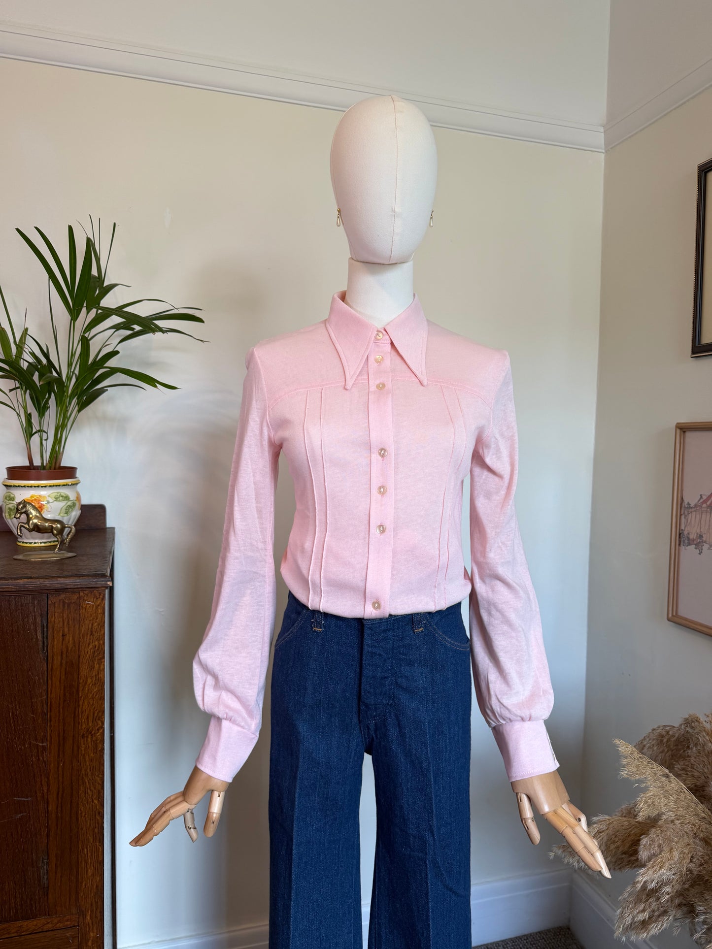 A pink long-sleeve button-up shirt with a pointed collar and front buttons, displayed on a mannequin torso with dark blue jeans in an indoor room setting.