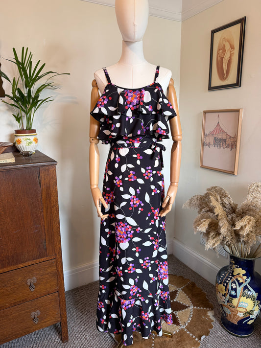 Vintage 1970s black maxi dress with thin straps, a tiered ruffle bodice and matching waist tie. The dress features a bold floral and berry print in shades of pink, purple and ivory with a softly flared hem, displayed on a mannequin in a vintage-inspired interior.