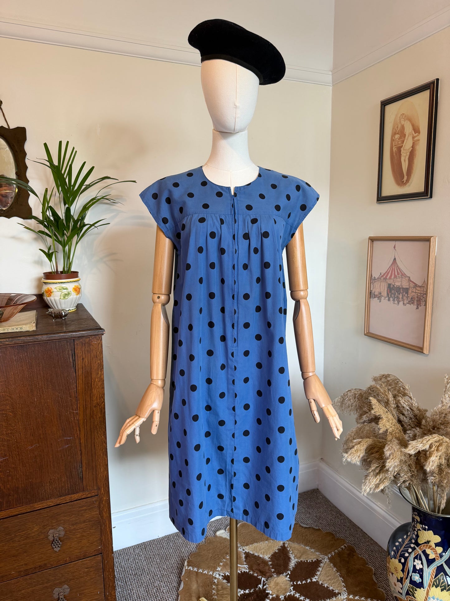 Vintage 1970s cornflower blue day dress with black polka dots, cap sleeves and a relaxed gathered shift silhouette. The dress features concealed front buttons and is displayed on a mannequin styled with a black beret in a vintage-inspired interior.