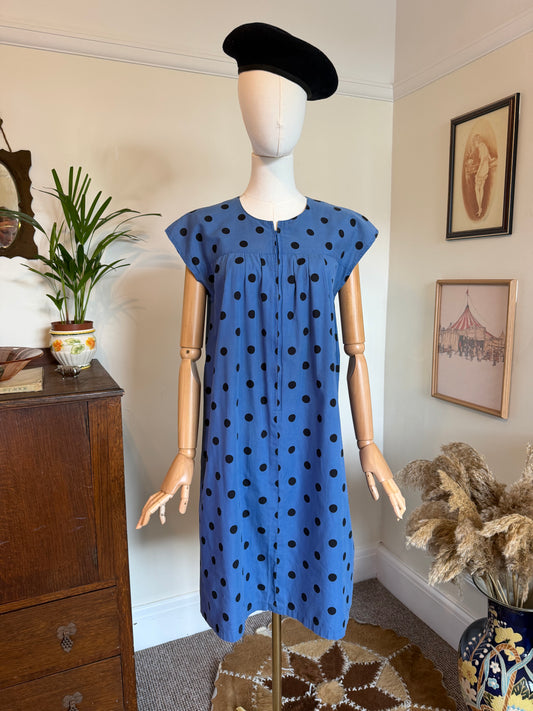 Vintage 1970s cornflower blue day dress with black polka dots, cap sleeves and a relaxed gathered shift silhouette. The dress features concealed front buttons and is displayed on a mannequin styled with a black beret in a vintage-inspired interior.