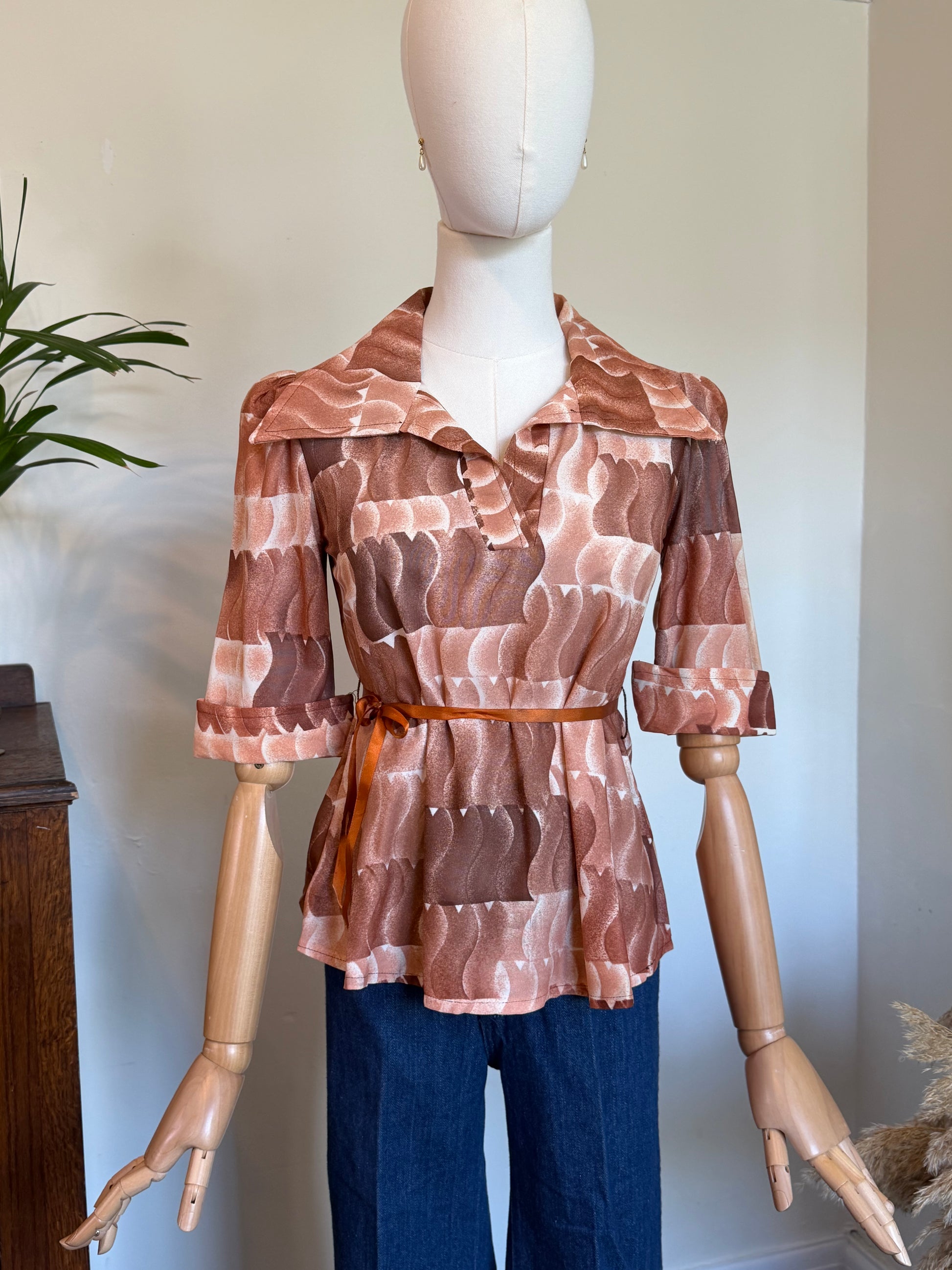 A vintage short-sleeve peplum blouse with a wide collar in an abstract wavy pattern in shades of brown, terracotta, and beige, styled on a wooden mannequin with blue jeans.