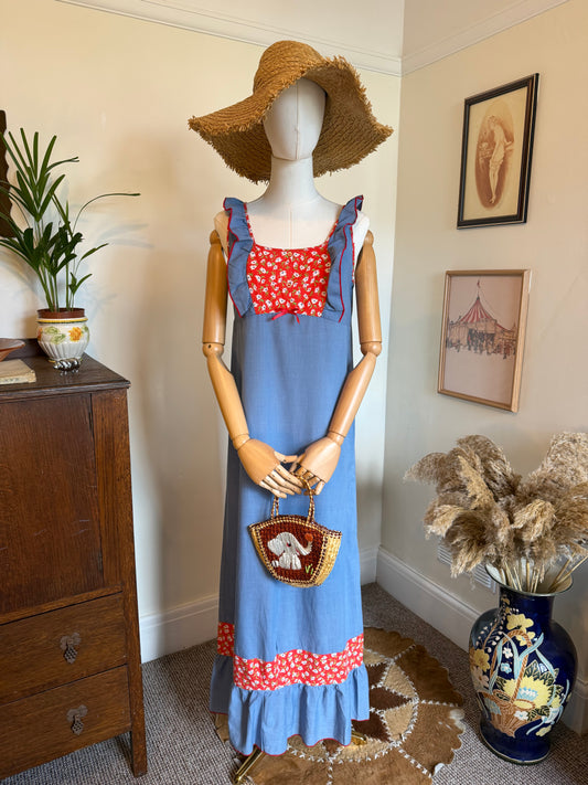 Handmade 1970s maxi dress in soft chambray blue with red and white floral contrast panels at the bodice and hem. Features ruffled shoulder straps with red piping, a square neckline and gathered ruffle hem. Displayed on a mannequin styled with a large straw sunhat and a small woven handbag featuring a white elephant motif.