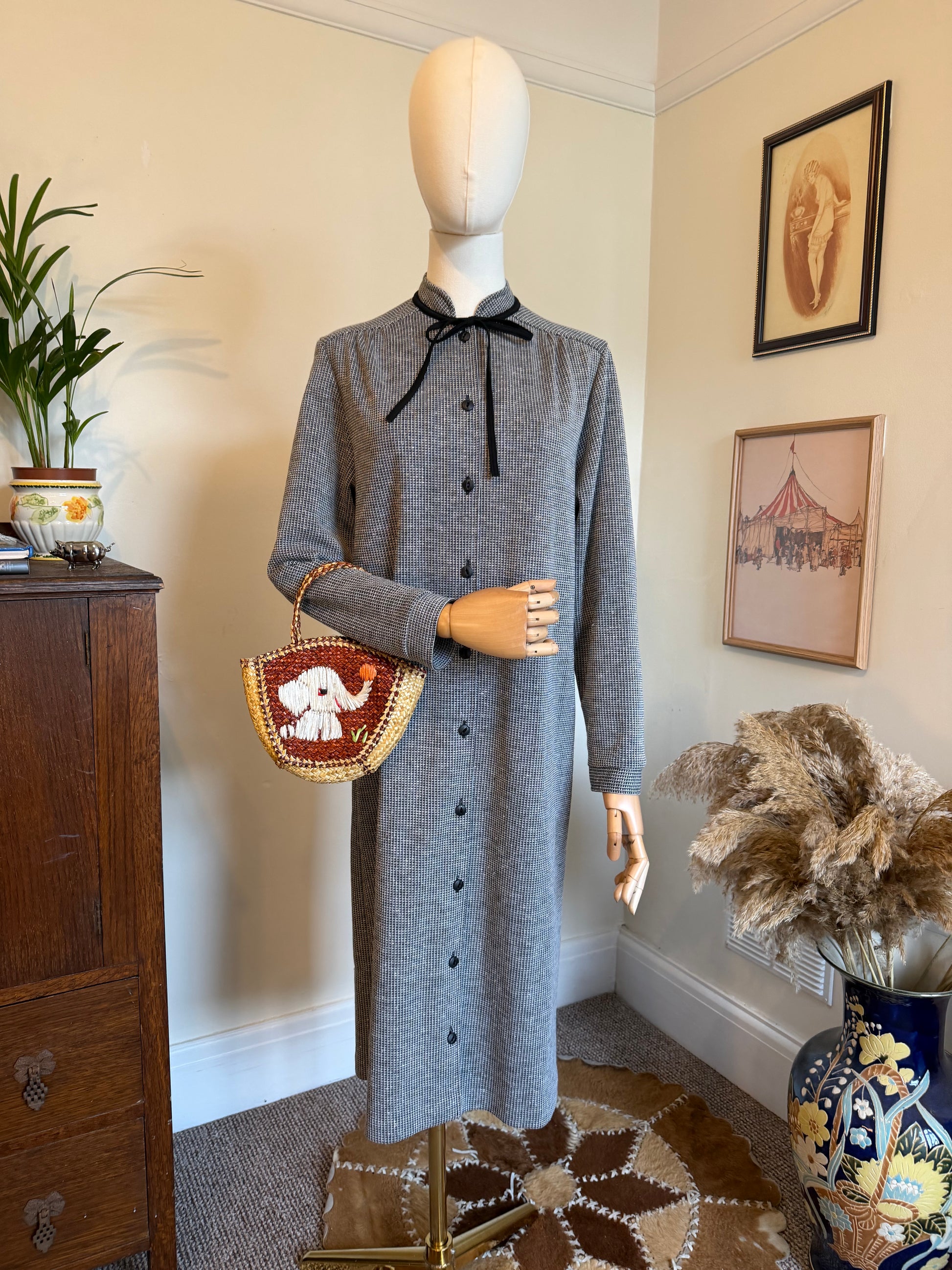 Vintage 1970s black and white micro-check midi dress with a button-through front, long sleeves and black ribbon tie-neck detail, displayed on a mannequin holding a woven handbag with an elephant motif.