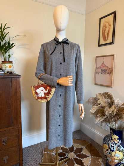 Vintage 1970s black and white micro-check midi dress with a button-through front, long sleeves and black ribbon tie-neck detail, displayed on a mannequin holding a woven handbag with an elephant motif.