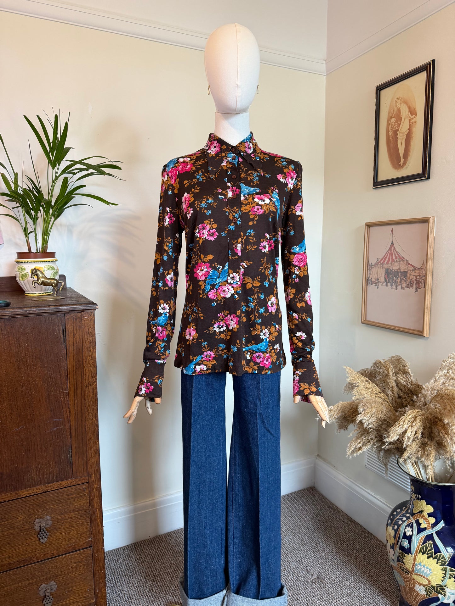 A long-sleeve button-up shirt with a colorful floral print on a dark brown base, styled on a mannequin with blue wide-leg jeans in a home interior setting.