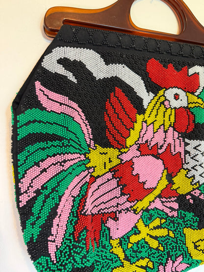 A close-up of a black beaded handbag featuring a colorful embroidered rooster design in pink, red, green, yellow, and white, with a brown wooden handle visible at the top.
