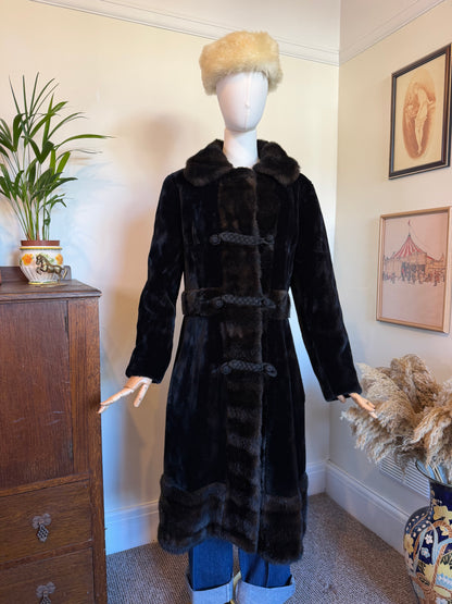 A long black fur coat with a plush collar and braided toggle closures, displayed on a mannequin in an indoor room. The mannequin also wears a beige fur hat and blue jeans, with a wooden cabinet, framed wall art, and pampas grass visible in the background.