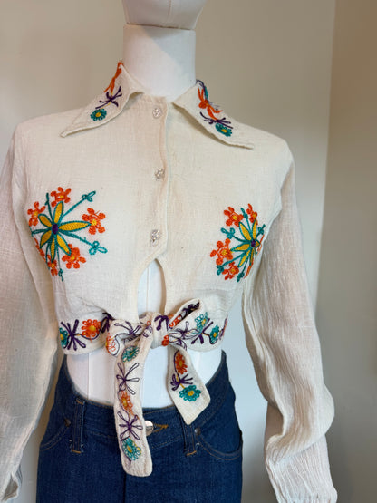 A cream-colored vintage 1970s long-sleeve button-up shirt with colourful floral embroidery on the front and collar, styled on a mannequin with blue jeans.