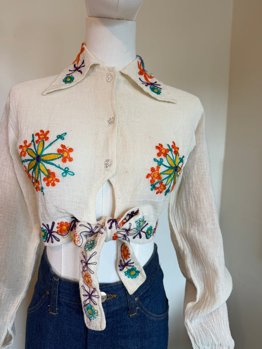A cream-colored vintage 1970s long-sleeve button-up shirt with colourful floral embroidery on the front and collar, styled on a mannequin with blue jeans.
