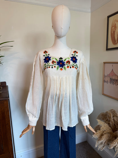 A cream-colored long-sleeve blouse with colorful floral embroidery across the chest, displayed on a mannequin with blue jeans in an indoor room setting.
