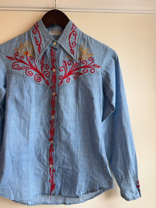 A light blue denim western-style button-up shirt hanging on a dark wooden hanger against a plain beige wall. The shirt features red embroidered scrollwork and floral patterns on the chest, along with a vertical red embroidered stripe down the center front placket.