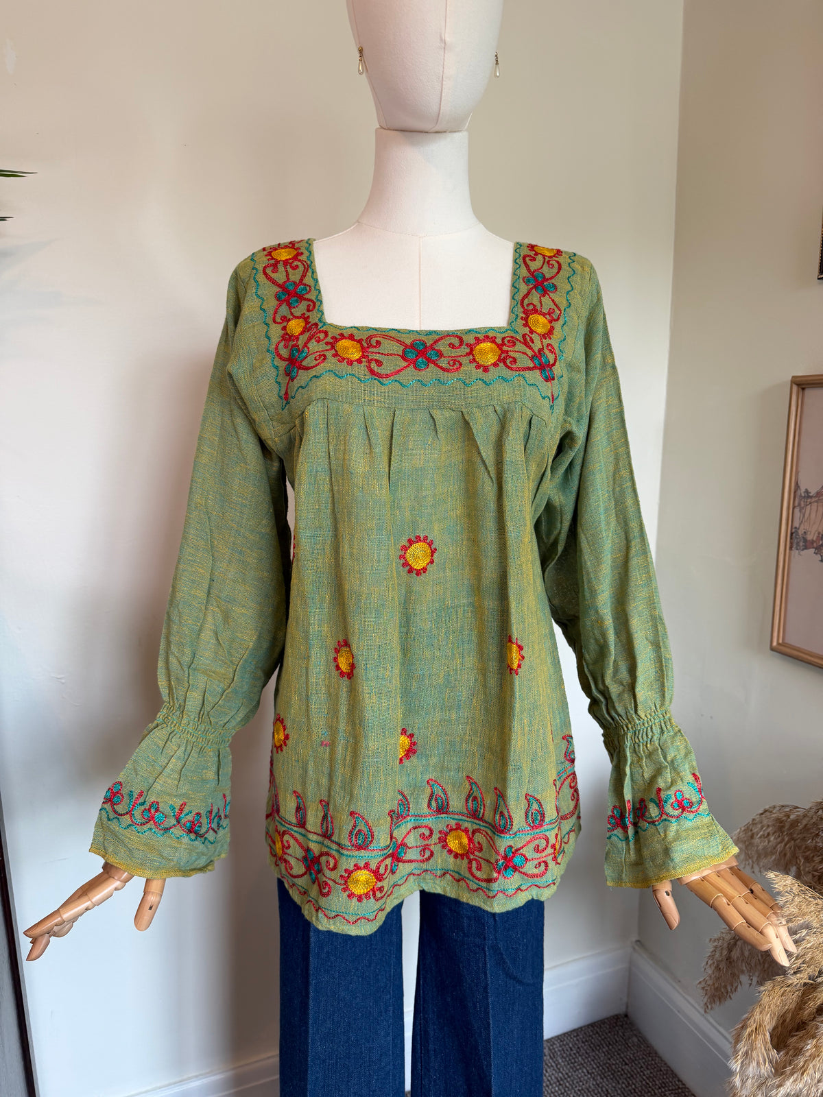 A green bohemian-style embroidered blouse displayed on a mannequin with blue jeans. The blouse features a square neckline with colorful floral embroidery in red, orange, and yellow, and matching embroidered cuffs.