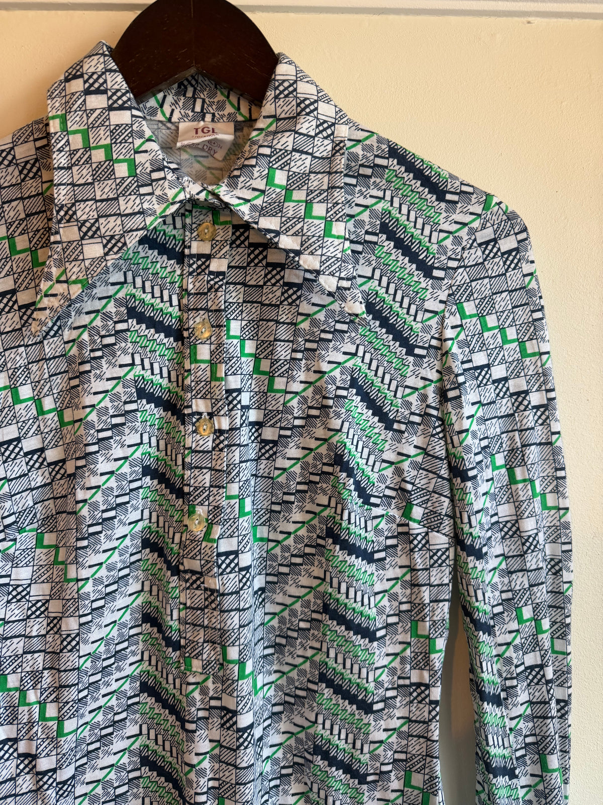 A long-sleeve vintage button-up popover shirt with a geometric pattern in green, navy, black, and white, hanging on a dark wooden hanger against a light beige wall.