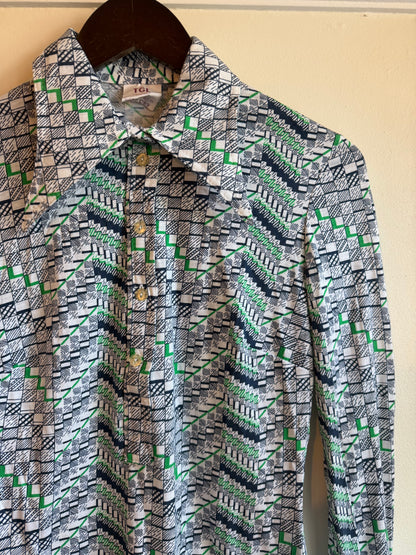 A long-sleeve vintage button-up popover shirt with a geometric pattern in green, navy, black, and white, hanging on a dark wooden hanger against a light beige wall.