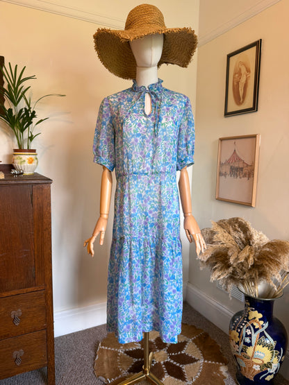 Vintage 1970s pastel blue floral midi dress with a lilac and green wildflower print, ruffle neckline, keyhole tie detail, puff sleeves, dropped waist and tiered skirt, displayed on a mannequin wearing a wide-brim straw hat.